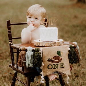 May include: A baby boy sits in a wooden high chair with a white cake decorated with a gold glitter "One" topper. A burlap banner with a duck design and the word "ONE" is draped over the chair. The banner has brown, green, and blue tassels.