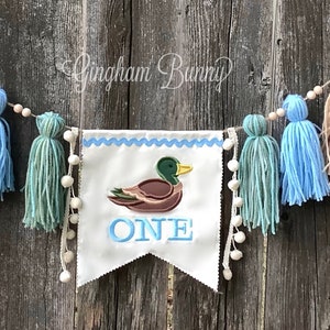 May include: A white banner with blue trim and the word "ONE" embroidered in blue. A brown duck with a green head is embroidered above the word "ONE". The banner is strung with a beige twine and decorated with beige, blue, and green yarn tassels.