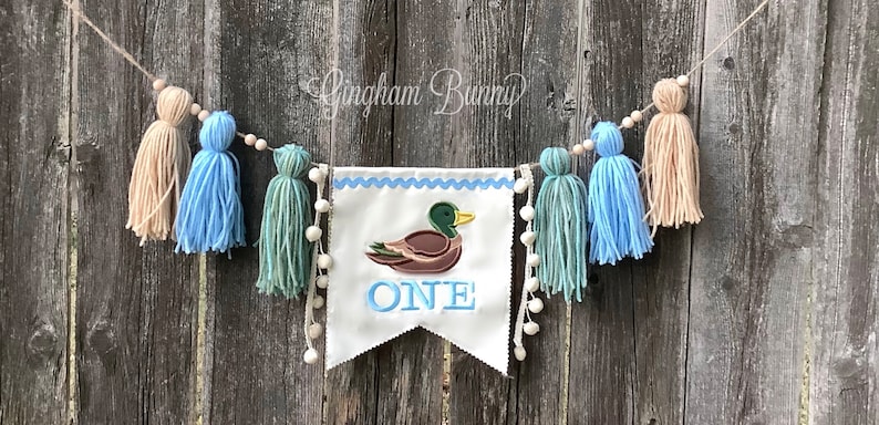 May include: A white banner with blue trim and the word "ONE" embroidered in blue. A brown duck with a green head is embroidered above the word "ONE". The banner is strung with a beige twine and decorated with beige, blue, and green yarn tassels.