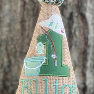 May include: A burlap-textured birthday party hat with a fishing theme. The hat features a green number 1, a fishing rod, a bucket, and the name "Elliot" in teal. A white-trimmed edge and a fluffy blue and green pom-pom top the hat.