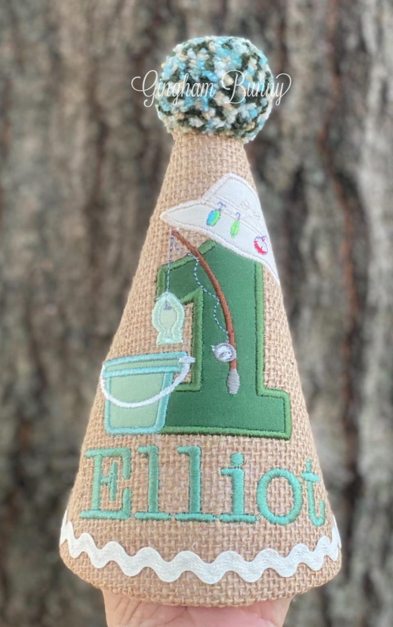 May include: A burlap-textured birthday party hat with a fishing theme. The hat features a green number 1, a fishing rod, a bucket, and the name "Elliot" in teal. A white-trimmed edge and a fluffy blue and green pom-pom top the hat.