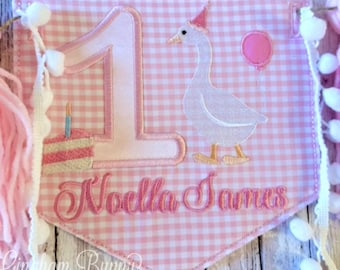 May include: Pink and white gingham fabric banner with a white goose holding a pink balloon, a number one, a pink cake with a candle, and the name "Noella James" embroidered in pink.