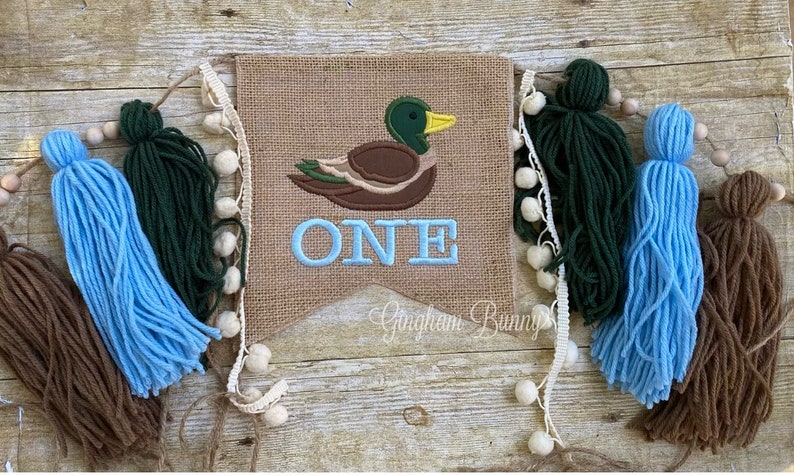 May include: A burlap banner featuring a brown duck and the word "ONE" embroidered in blue. The banner is adorned with brown, blue and green tassels.