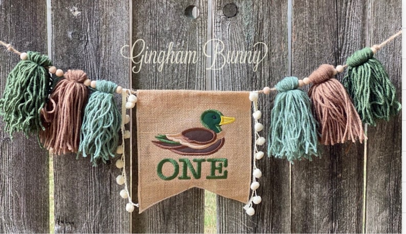 May include: A burlap banner featuring an embroidered duck in shades of green, brown and yellow, with the word "ONE" in green. The banner is strung with brown wooden beads and green and brown yarn tassels.