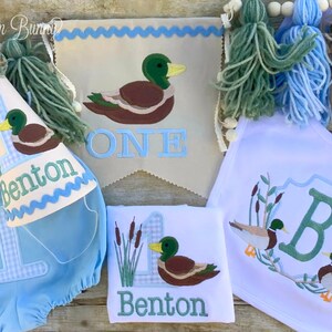 Puede incluir: Un conjunto de fiesta de primer cumplea&ntilde;os para beb&eacute; con tem&aacute;tica de pato. El conjunto incluye un pa&ntilde;al de gingham azul y blanco, un babero blanco con una letra B verde y patos, un body blanco con un pato y el nombre Benton, una pancarta blanca con la palabra "ONE" y un pato, y un gorro de gingham azul y blanco con un pato y el nombre Benton.