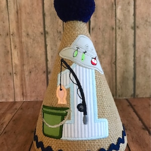 May include: A burlap cone-shaped party hat with a blue pom-pom on top. The hat is decorated with a blue and white striped fabric, a green bucket, a fishing rod, a fish, and a number one. The hat has a blue and white scalloped trim around the bottom.