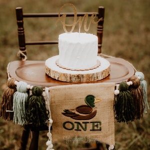 May include: A white cake with a gold glitter "One" topper sits on a wooden slice. A burlap banner with a green embroidered duck and the word "ONE" hangs below the cake. The banner also says "Gingham Bunny".