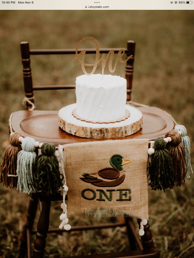 May include: A white cake with a gold glitter "One" topper sits on a wooden slice. A burlap banner with a green embroidered duck and the word "ONE" hangs below the cake. The banner also says "Gingham Bunny".