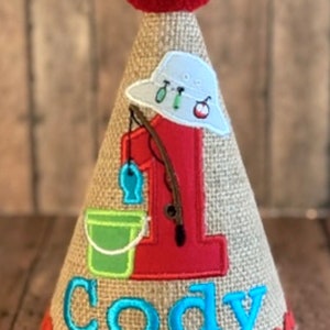 May include: A burlap-textured birthday party hat with a red pom-pom on top. The hat features a red number 1, a fishing-themed design, and the name "Cody" in turquoise. A red zigzag trim accents the base.