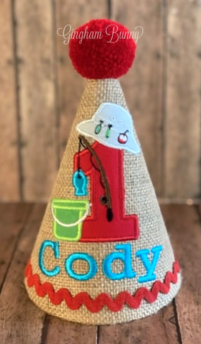 May include: A burlap-textured birthday party hat with a red pom-pom on top. The hat features a red number 1, a fishing-themed design, and the name "Cody" in turquoise. A red zigzag trim accents the base.