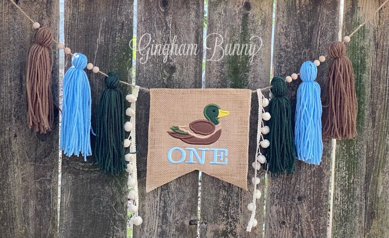 May include: A burlap banner featuring an embroidered duck and the word "ONE". The banner is adorned with blue, green and brown yarn tassels and white pom-poms.