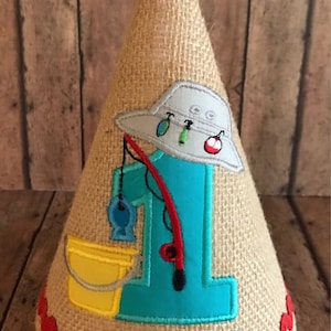 May include: A burlap cone-shaped party hat with a red and white bobber on top. The hat features a blue number 1, a fishing hat, a fishing rod, a yellow bucket, and a red trim. The hat is decorated with fishing-themed elements.