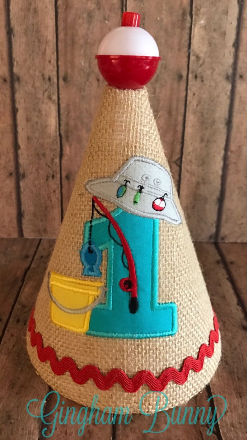 May include: A burlap cone-shaped party hat with a red and white bobber on top. The hat features a blue number 1, a fishing hat, a fishing rod, a yellow bucket, and a red trim. The hat is decorated with fishing-themed elements.