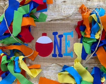 May include: A burlap banner with the word "ONE" in blue and turquoise letters, a red and white fishing bobber, and a string of colorful fabric scraps.