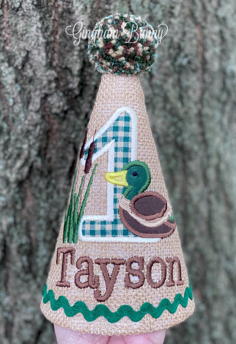 May include: A burlap birthday hat with a green and white gingham number one, a green and brown embroidered duck, and the name "Tayson" embroidered in brown. The hat has a green and white pom-pom on top.