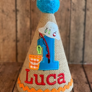 May include: A burlap cone-shaped birthday hat with a blue pom-pom on top. The hat features a blue number 1 with a fishing theme, including a fishing rod, fish, and a bucket. The name "Luca" is embroidered in red.