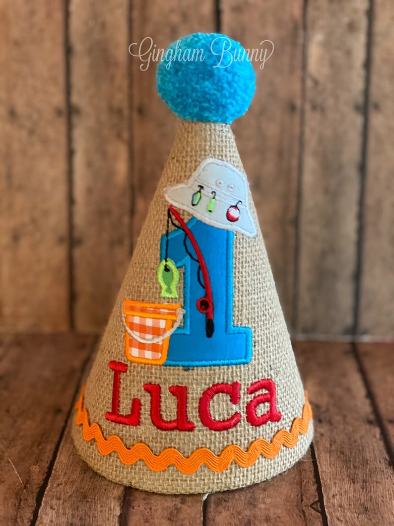 May include: A burlap cone-shaped birthday hat with a blue pom-pom on top. The hat features a blue number 1 with a fishing theme, including a fishing rod, fish, and a bucket. The name "Luca" is embroidered in red.