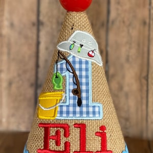 May include: A burlap cone-shaped birthday hat with a fishing theme. It features a red and white bobber, a blue gingham "1", a fishing rod, and the name "Eli" in red. A yellow bucket and a blue wave trim complete the design.