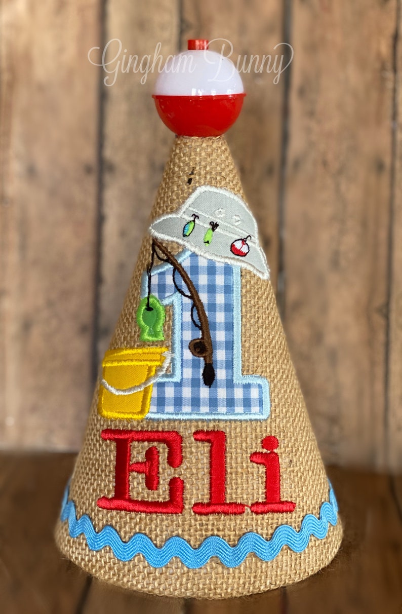May include: A burlap cone-shaped birthday hat with a fishing theme. It features a red and white bobber, a blue gingham "1", a fishing rod, and the name "Eli" in red. A yellow bucket and a blue wave trim complete the design.