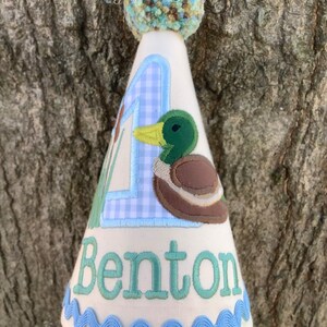 Puede incluir: Un gorro de fiesta de cumplea&ntilde;os blanco con un n&uacute;mero uno de vichy azul y blanco, un pato bordado en verde y marr&oacute;n, y el nombre "Benton" bordado en verde. El gorro tiene un pomp&oacute;n azul y blanco en la parte superior.