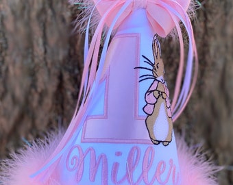 May include: A pink and white birthday party hat with a pink bow, a white number one, and a Peter Rabbit character. The hat is trimmed with pink feathers and the name "Miller" is embroidered on the front. The hat also has the words "Gingham Bunny" on the bottom.