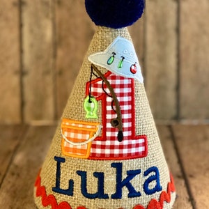 May include: A burlap-textured birthday party hat with a fishing theme. The hat features a blue pom-pom, a red and white gingham number one, and the name "Luka" in blue. It is decorated with fishing-related appliqu&eacute;s.