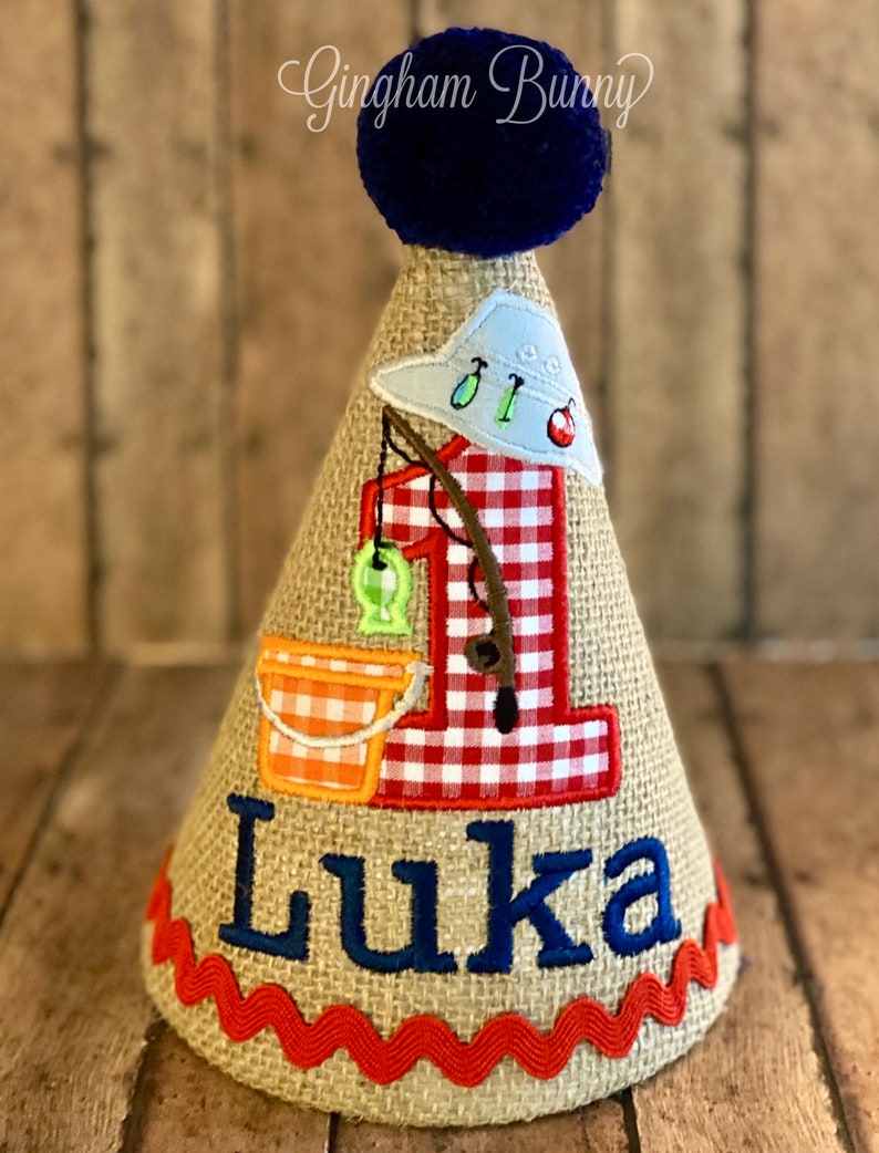 May include: A burlap-textured birthday party hat with a fishing theme. The hat features a blue pom-pom, a red and white gingham number one, and the name "Luka" in blue. It is decorated with fishing-related appliqu&eacute;s.