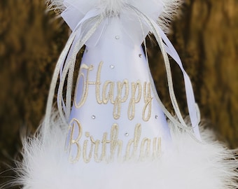 May include: White birthday party hat with a fluffy feather trim and a large white pom-pom on top. The hat has the words "Happy Birthday" in gold script and is decorated with small rhinestones.