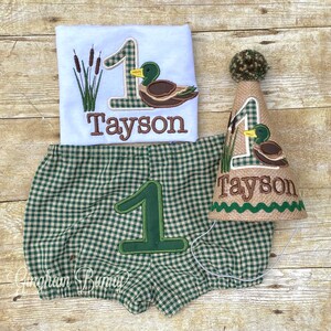 May include: A green and white checked baby outfit with a brown duck and the number one embroidered on it. The outfit includes a shirt, nappy cover, and a party hat. The name "Tayson" is embroidered on the shirt and hat.