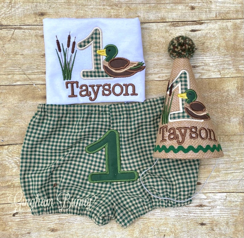 May include: A green and white checked baby outfit with a brown duck and the number one embroidered on it. The outfit includes a shirt, nappy cover, and a party hat. The name "Tayson" is embroidered on the shirt and hat.