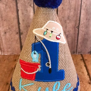 May include: A burlap birthday party hat with a blue pom-pom on top. The hat is decorated with a blue number one, a fishing rod, a bucket, and a fishing hat. The name "Kyle" is embroidered on the hat in blue thread.