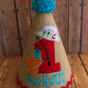 May include: A burlap cone-shaped birthday hat with a blue pom-pom on top. The hat features a red number 1, a fishing-themed design, and the name "Beckett" embroidered in blue. A red, wavy trim adorns the base.