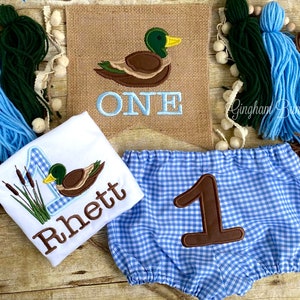 May include: A baby boy's first birthday outfit featuring a duck and reeds design on a white onesie, blue and white gingham bloomers with a brown number one, and a burlap banner with a duck and the word "ONE" in blue.