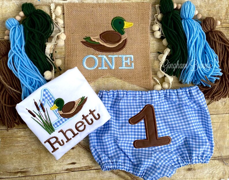 May include: A baby boy's first birthday outfit featuring a duck and reeds design on a white onesie, blue and white gingham bloomers with a brown number one, and a burlap banner with a duck and the word "ONE" in blue.