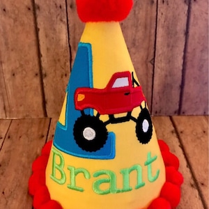 May include: Yellow birthday party hat with a red pom-pom on top. The hat has a blue and yellow number one, a red monster truck, and the name "Brant" embroidered on it. The hat is decorated with a red pom-pom trim.