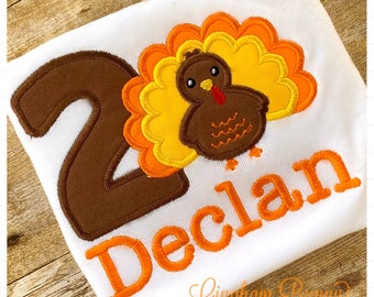 May include: A white t-shirt with an embroidered brown number "2" and a brown turkey with orange and yellow feathers. The name "Declan" is embroidered in orange below the turkey.