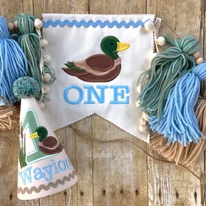 Puede incluir: Una pancarta azul y blanca con un pato y la palabra "ONE" bordada. Un gorro de fiesta azul y blanco con un pato y el nombre "Waylon" bordado. La pancarta y el gorro est&aacute;n decorados con borlas azules, verdes y marrones.