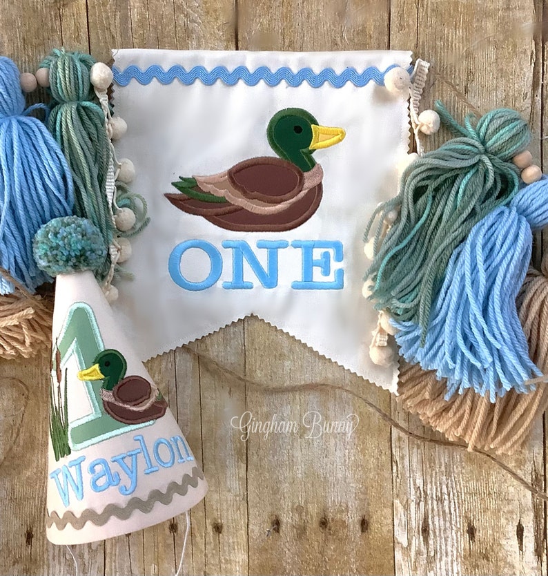 Puede incluir: Una pancarta azul y blanca con un pato y la palabra "ONE" bordada. Un gorro de fiesta azul y blanco con un pato y el nombre "Waylon" bordado. La pancarta y el gorro est&aacute;n decorados con borlas azules, verdes y marrones.
