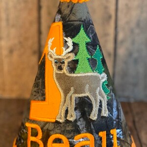 May include: A camouflage fabric party hat with an orange pom-pom on top. The hat has an orange number one, a brown deer, and green trees embroidered on it. The name "Beau" is embroidered in orange on the bottom of the hat.