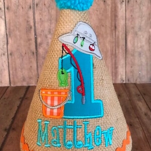 May include: A burlap-textured birthday party hat with a blue pom-pom on top. The hat features a fishing theme with a blue number 1, a fishing rod, a bucket, and the name "Matthew" in turquoise. The hat is trimmed with orange.
