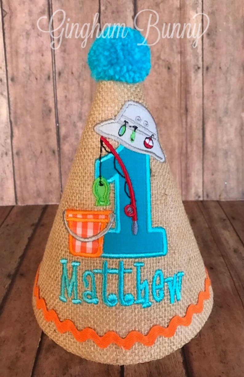 May include: A burlap-textured birthday party hat with a blue pom-pom on top. The hat features a fishing theme with a blue number 1, a fishing rod, a bucket, and the name "Matthew" in turquoise. The hat is trimmed with orange.