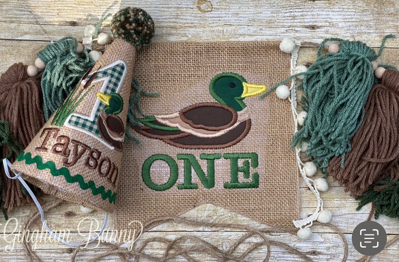May include: A burlap banner featuring an embroidered green and brown duck and the word "ONE" in green. A matching burlap party hat with a green and white gingham number one, a small embroidered duck, and the name "Tayson" is also included.