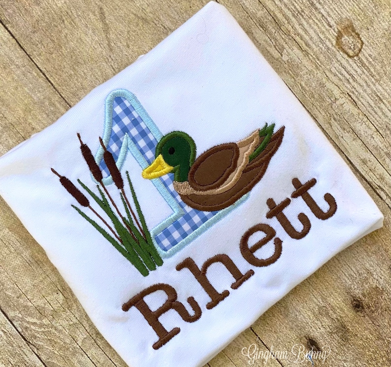 May include: A white onesie with a blue and white gingham number one, a brown duck with green head and yellow beak, and green reeds. The name "Rhett" is embroidered in brown below the duck.
