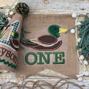 May include: A burlap banner with a green and brown embroidered duck and the word "ONE" in green. A matching burlap party hat with a green and white gingham number one, a small embroidered duck, and the name "Tayson" is also included.