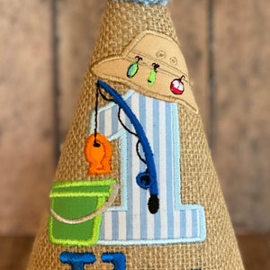 May include: A burlap cone-shaped birthday hat with a blue pom-pom on top. The hat features an embroidered fishing theme with a fishing rod, fish, and the name "Ike" in blue. A blue wave trim accents the base.