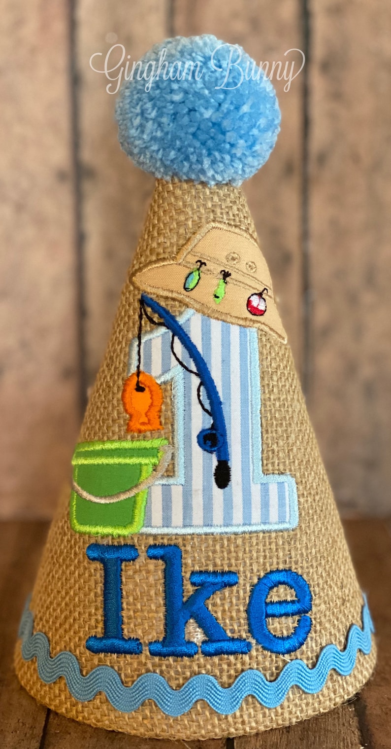 May include: A burlap cone-shaped birthday hat with a blue pom-pom on top. The hat features an embroidered fishing theme with a fishing rod, fish, and the name "Ike" in blue. A blue wave trim accents the base.
