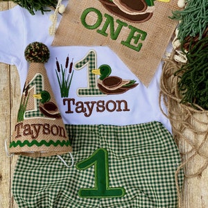 May include: A green and brown gingham outfit for a baby's first birthday. The outfit includes a hat, a onesie, and bloomers. The onesie has a number one and the name "Tayson" embroidered on it. The hat has a number one and a duck embroidered on it. The bloomers have a number one embroidered on them.