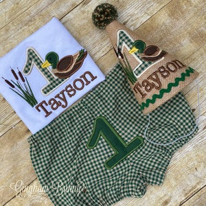 May include: A green and brown plaid baby outfit with a duck and the number one embroidered on it. The outfit includes a shirt, shorts, and a hat. The hat is brown with a green and white plaid pattern and has a green and brown duck embroidered on it. The hat also has the name "Tayson" embroidered on it.