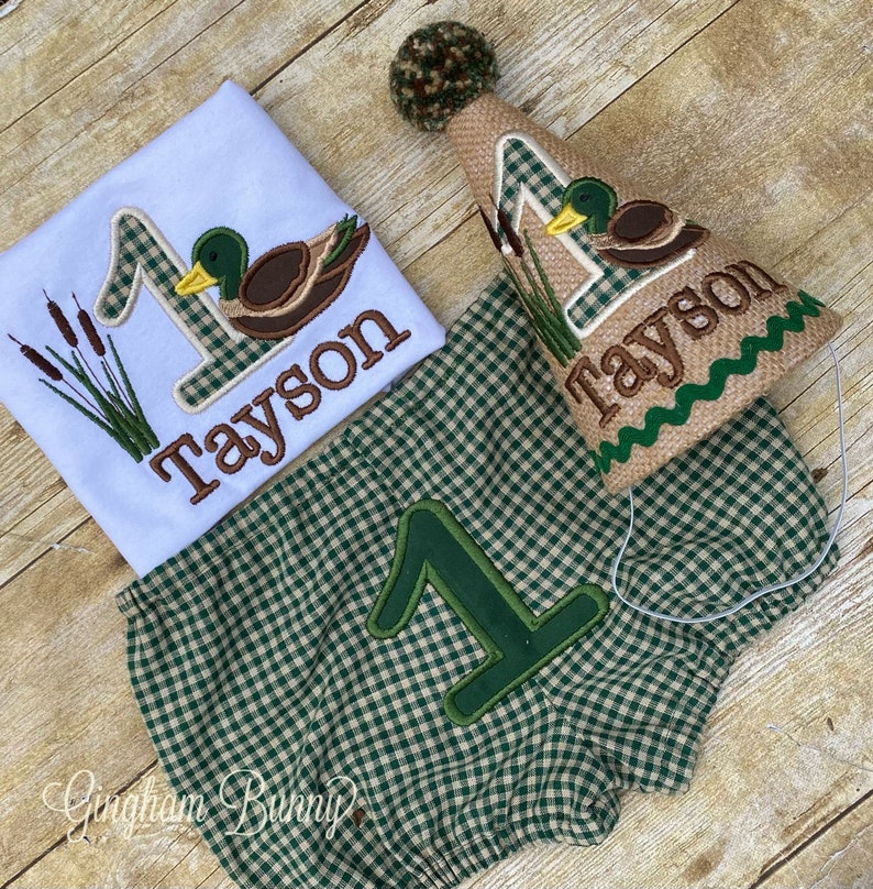 May include: A green and brown plaid baby outfit with a duck and the number one embroidered on it. The outfit includes a shirt, shorts, and a hat. The hat is brown with a green and white plaid pattern and has a green and brown duck embroidered on it. The hat also has the name "Tayson" embroidered on it.