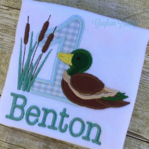 Puede incluir: Un pelele blanco con tela vichy azul y blanca, un pato verde y juncos verdes. El n&uacute;mero uno est&aacute; bordado en el centro del pelele. El nombre Benton est&aacute; bordado debajo del pato.